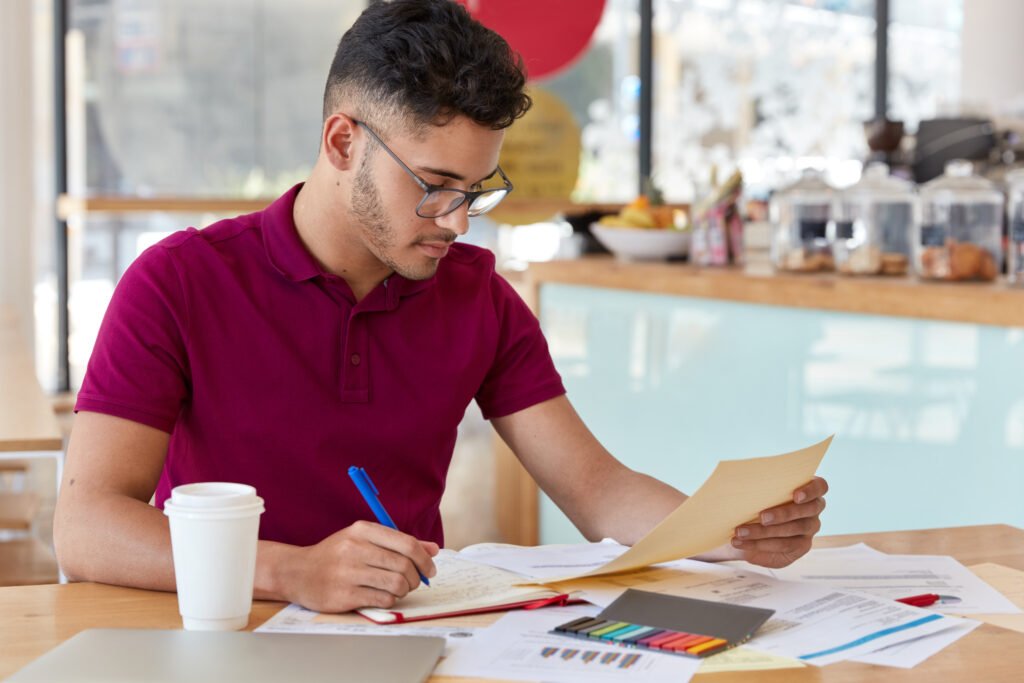 image of busy unshaven copy writer or college student dressed in casual clothing, makes notes in notebook, focused into document, looks attentively, poses at small cafetiera, drinks hot beverage.
